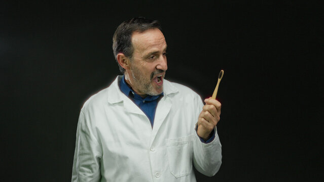 Man in doctor coat holding bamboo toothbrush against black background expresses surprise with hand gestures.