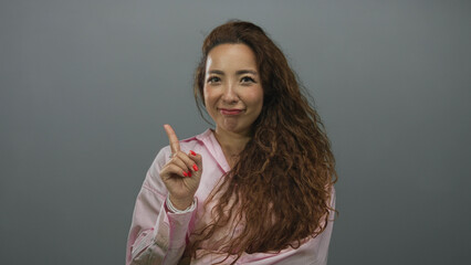 Woman points finger to chest wearing pink shirt in gray studio with long curly hair; skeptical doubt.