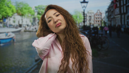 Fototapeta premium Woman with hand on neck on amsterdam street by canal boats and bikes, showing neck pain; discomfort.
