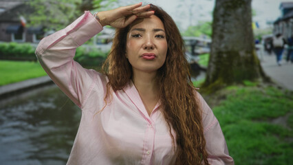 Woman with long curly hair and pink shirt holding hand to forehead shading eyes while smiling...