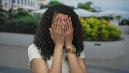 Woman covers face with hands on city street before raising both hands in playful reveal; joy surprise.