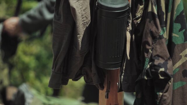 Close up view of military gear and apparel including a metal gas mask container, wool field cap, and patterned camouflage jacket hanging outdoors on a wooden post in Military field camp.