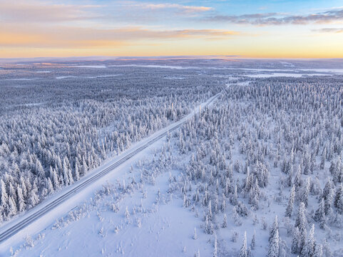 Aerial view of a ribbon of road cuts through a pristine, snow-laden forest under a pastel dawn sky, Lapland, Finland.
