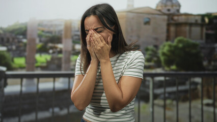 Fototapeta premium Woman holds head with hands to ears, wincing as a visible headache while standing on a stone balcony overlooking ancient ruins and an old building; distress.