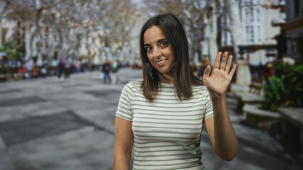 Fototapeta premium Woman hispanic young waving right hand and smiling toward camera on a pedestrian city street lined with trees, benches and blurred passersby; friendly greeting.