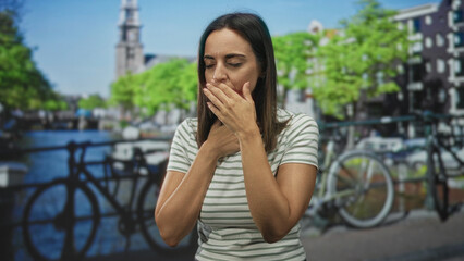 Fototapeta premium Woman young hispanic holding throat with both hands beside a parked bicycle on a street by a canal and bridge; breathing difficulty anxiety.