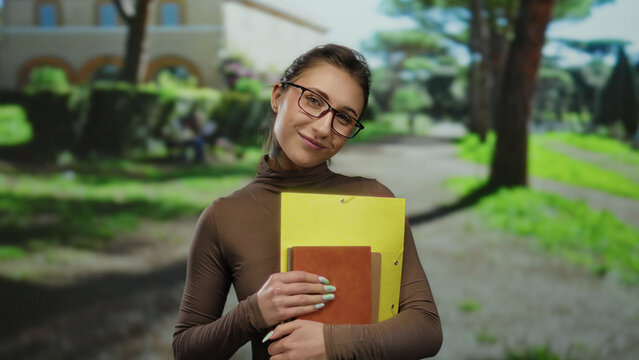 Young woman with glasses holding folders smiling in a sunlit park setting showcasing joy creativity and learning outdoors surrounded by lush greenery and a clear blue sky.