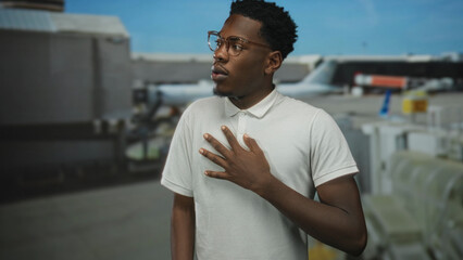 Fototapeta premium Man hand on chest stands at airport terminal outdoors by parked plane under bright blue sky; surprise astonishment wonder shock.