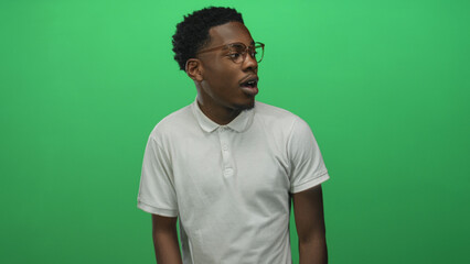 Young african american man wearing white polo shirt with glasses turns head in bright green studio;...