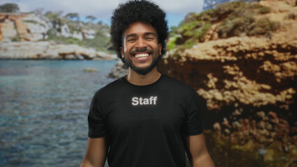 Man smiling in staff uniform at picturesque seaside location with rocky cliffs and clear blue water, exuding a cheerful and friendly atmosphere outdoors.