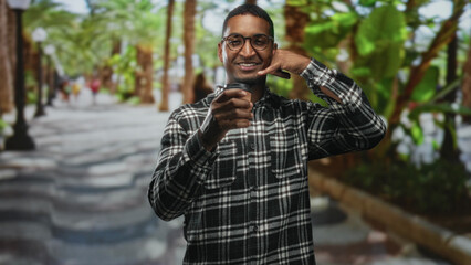 Fototapeta premium Young black man holding a takeaway coffee cup and making a call me gesture on a tree lined street; friendly greeting.