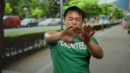 Man wearing green volunteer shirt on urban street with blurred cars and trees in background, expressing energetic gesture and youthful vigor.