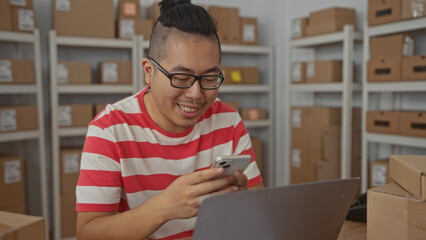 Man wearing glasses holding smartphone and typing with both hands beside laptop amid stacked parcel boxes on shelves in an office building, smiling; small business productivity.