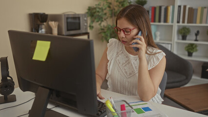 Woman holds phone to ear while reviewing printed charts and looking at computer monitor at office building desk; concentration productivity.
