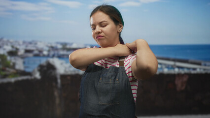 Young caucasian woman holding shoulder at seaside building wearing apron and striped tshirt near boats and harbor; pain exhaustion.