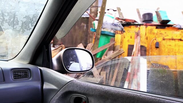 Perspective from inside a vehicle, passenger side window and side mirror reveal water drops on glass. Outside view features a cluttered, rundown urban environment with makeshift structures