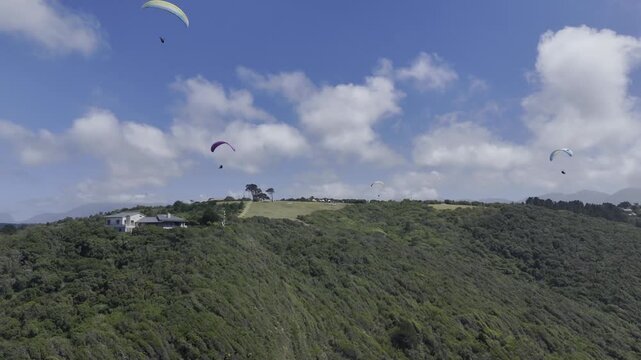 Drone flies to the right as paragliders fill the partially cloudy sky on a sunny day on the Garden Route in Wilderness, South Africa