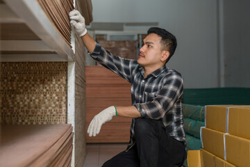 Asian adult male carpenter reaching upward to inspect stacked wood panels on upper shelf, woodworking business owner managing material condition and inventory inside interior furniture material store