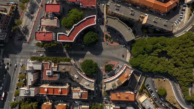 Top-down aerial view of a symmetrical urban plaza. Zenithal shot of curving residential buildings with red roofs frame a central street, showcasing unique rationalist architecture and city planning.