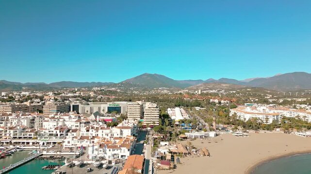Aerial tracking back over Puerto Ban&uacute;s marina and beach in Marbella, Southern Spain, and La Concha mountains under sunny blue skies.