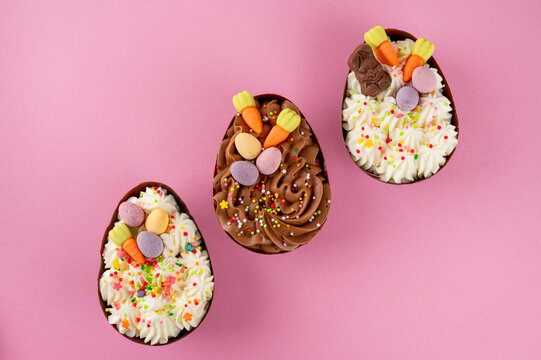 Easter dessert. Chocolate eggs with cheesecake filling decorated with chocolate candies and sugar sprinkles on a pink background. Copy space, top view.