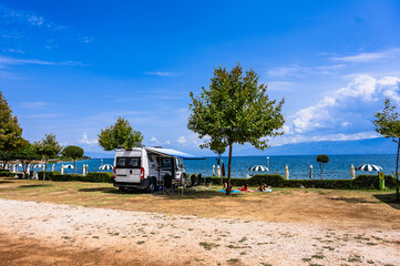 A scenic summer day at a Lake Ohrid campsite in Udenisht, Albania, featuring a camper van with an awning and a family sunbathing on the grass under a tree with clear blue waters in the background.