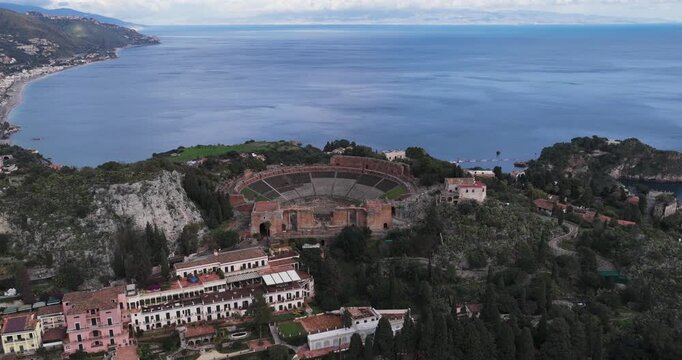 Aerial view of the Teatro Antico di Taormina, with its ancient stone structure contrasting against the deep blue sea, Taormina, Sicily, Italy.