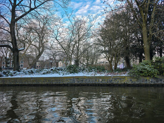 Snowy Amsterdam park next to canal in winter: bare trees, light dusting of white snow on ground, tranquil cold season urban nature scene, January 2026