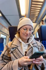 Portrait young adult woman passenger use smartphone inside modern train wear winter hat glasses coat commuting on sunny day. Casual travel lifestyle scene public transport interior urban routine