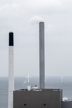 Industrial coastal infrastructure with chimney stack and wind turbine showing energy transition in haze above copenhagen waterfront skyline