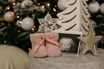 Small knitted pink baby shoes stand next to Christmas toys on the background of a Christmas tree in a photo studio