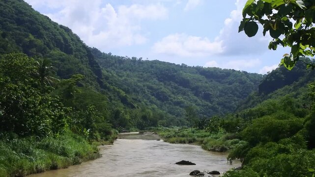 View of the Oyo River in Kedungjati, Selopamioro, Imogiri, Yogyakarta, Indonesia, flanked by two hills with lush green plants growing around it with a bright blue sky in the background during the day