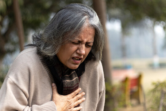 Indian senior woman holding chest with pained expression sitting on bench outdoors. Possible heart diseases problem, outdoor got troubles with her chest pain or breathing difficulty in park