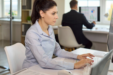 Woman employee in office typing finance data. Focused analyst works at computer with spreadsheets, preparing report while a colleague works nearby. Concept of professional financial workflow.
