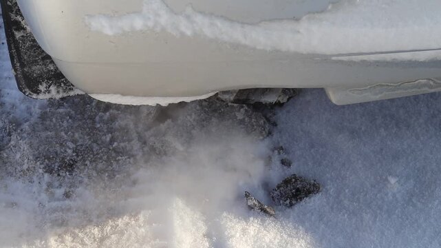 Snow piles up around the exhaust pipe of a parked car during winter. Cold air creates a cloud of exhaust as the engine runs. Snow covers the ground.