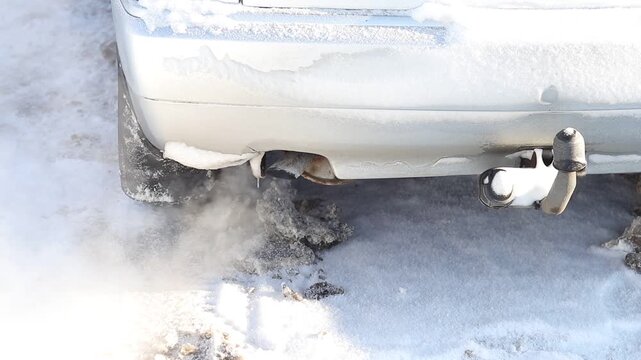 Snow piles up around the exhaust pipe of a parked car during winter. Cold air creates a cloud of exhaust as the engine runs. Snow covers the ground.