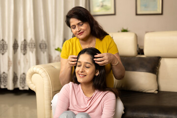 Fototapeta premium Indian happy mother combing young daughter's hair in living room when sitting on sofa at home together.