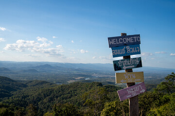 Wooden welcome sign at a scenic viewpoint overlooking green mountains and valleys in northern Thailand. Clear blue sky and layered hills create a peaceful travel and nature landscape.