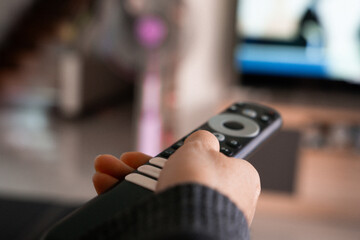 Close-up of a hand holding a television remote control with blurred living room and TV screen in the background. Concept of home entertainment, technology use, and modern lifestyle.