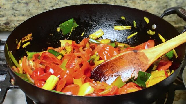 close-up chopped red bell peppers, green onions, and sweet chili peppers being saut&eacute;ed in dark iron pot. Wooden spatula is placed among colorful vegetables on kitchen stove
