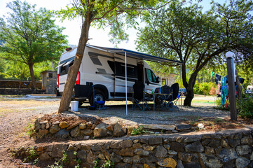 Camper van set up at a shaded campsite near Komani Lake in Albania, with chairs and awning among trees, capturing peaceful lakeside camping and the freedom of van life in nature.