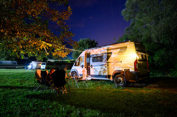 Night camping in nature near river Zeta, Montenegro, with a camper van, chairs, and warm lights under trees, capturing a cozy riverside camping atmosphere and van life experience.