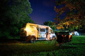 Night camping in nature near river Zeta, Montenegro, with a camper van, chairs, and warm lights under trees, capturing a cozy riverside camping atmosphere and van life experience.