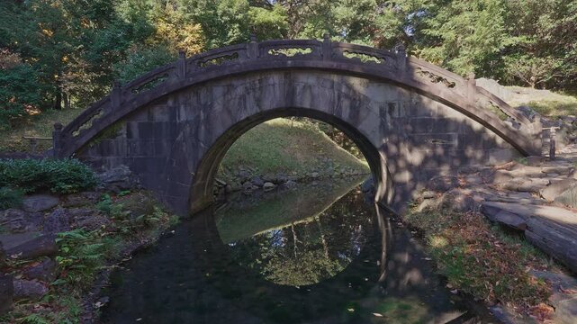 A tranquil view of the historic Engetsu-kyo (Full Moon Bridge) stone arch bridge, where the reflection in the still water creates a perfect circular "full moon" effect, framed by lush forest greenery.