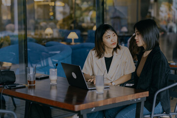 Two young asian businesswoman working and discussing their project together while using digital devices a laptop and mobile phone in cafe.