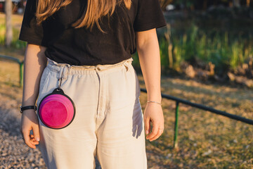 A young woman with long brown hair wears a black t-shirt and light-colored pants. A pink dog treat pouch hangs from her waist in a park setting.