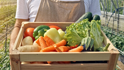A farmer holds a wooden crate of fresh vegetables—carrots, zucchini, lettuce, tomatoes—in a greenhouse, emphasizing sustainable agriculture and local, organic produce.