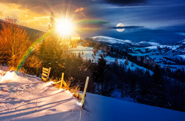 countryside in mountains. time change concept. lovely scenery with wooden fence and coniferous forest on snowy slope with sun and moon. village on distant hill. uncertainty, trust issue and relations
