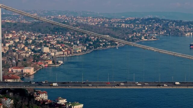 Aerial drone footage of the Istanbul Bosphorus, featuring the 15 July Martyrs Bridge, coastal neighborhoods, red-roof houses and wide blue waters, presenting a clear panoramic view of the city.