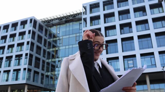 Student walking home reads review of her thesis holding papers and playfully shows horns gesture reacting to harsh criticism humorous frustrated moment near campus buildings